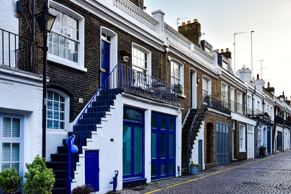 A row of multi-story terraced houses with dark brick facades and white window frames, situated along a cobblestone street. Each house has an outdoor staircase leading to a small balcony with metal railings, painted in blue. The ground floors feature large glass doors and windows, some with white shutters or window boxes with plants. A few potted plants are visible near the entrances. The image is taken during daylight, with soft natural lighting illuminating the buildings. This setting illustrates urban residential architecture typical of Hammersmith, where house removals may involve navigating narrow streets and stairs. The presence of gardening elements and the compact arrangement of the houses highlights common considerations in home relocation and furniture transport in such environments. Hammersmith Movers could assist with these logistics, ensuring smooth packing and loading processes for house removals in areas with limited street access.