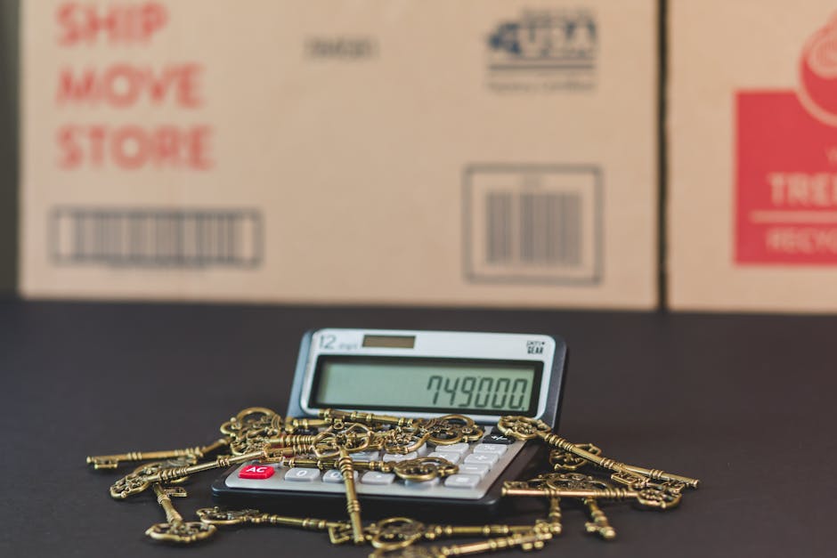 A close-up view of a calculator displaying the number 749800, surrounded by a pile of brass keys with various shapes and sizes, some attached to small chains. The items are placed on a black surface with cardboard boxes in the background, marked with red and black text and barcodes. The scene appears to be set indoors, possibly in a storage or moving environment, with the keys possibly representing valuables or items to be transported during a house removal. The background boxes suggest packing and relocation activities, and the presence of the calculator hints at cost or inventory calculations involved in the moving process, consistent with the services provided by Hammersmith Movers in home relocation and furniture transport.