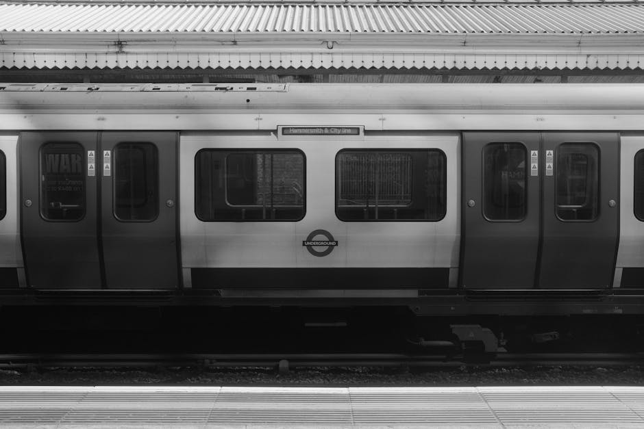A black and white image of a London Underground train carriage parked at an outdoor station platform. The carriage features multiple rectangular windows, some with protective grilles, and standard sliding doors. The platform edge with tactile paving strips is visible in the foreground, and a sign above indicates the station name 'Hammersmith & City'. The image captures the side view of the train, with the metallic surface reflecting minimal light. No people are visible, and the scene appears to be during a quiet period. This setting relates to house removals as part of transportation and logistics involved in home relocation, highlighting the importance of reliable transport services such as those provided by Hammersmith Movers for efficient furniture transport and moving processes.
