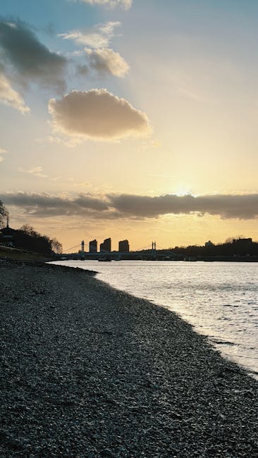The image shows a lakeside scene during sunset with a partly cloudy sky. In the foreground, a pebble beach extends along the water's edge, with small stones and gravel visible. To the left, a few trees with dark foliage are silhouetted against the sky. In the middle ground, several cardboard boxes, wrapped in plastic and secured with packing tape, along with a few pieces of furniture covered with fabric blankets, are arranged on the paved area next to a stationary moving van. The van is partially visible, with its rear doors open, revealing additional boxes and packing materials inside. The setting suggests a home relocation process with furniture and boxes ready for transport, possibly carried out by a moving company such as Hammersmith Movers, as indicated by the context of house removals and furniture transport. The scene captures the logistical aspect of packing and loading for a house move near the water during the late afternoon or early evening.