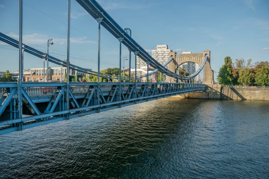 A clear view of the blue-painted Hammersmith Bridge spanning the River Thames in London during daytime, with the suspension cables and steel framework visible in the foreground. The bridge connects the Hammersmith area to nearby buildings, including modern apartments and historic structures, with trees showing early signs of autumn foliage on the riverbank. The water below reflects the bridge’s structure and the blue sky with a few scattered clouds. This setting illustrates the typical environment for house removals and furniture transport, where the bridge serves as a key route for moving trucks and vehicles involved in home relocation services. Occasionally, removals companies like Hammersmith Movers may operate in this area, coordinating loading and unloading of packed boxes, furniture covered with protective blankets, and large appliances at the start or end of the loading process as part of professional moving logistics.