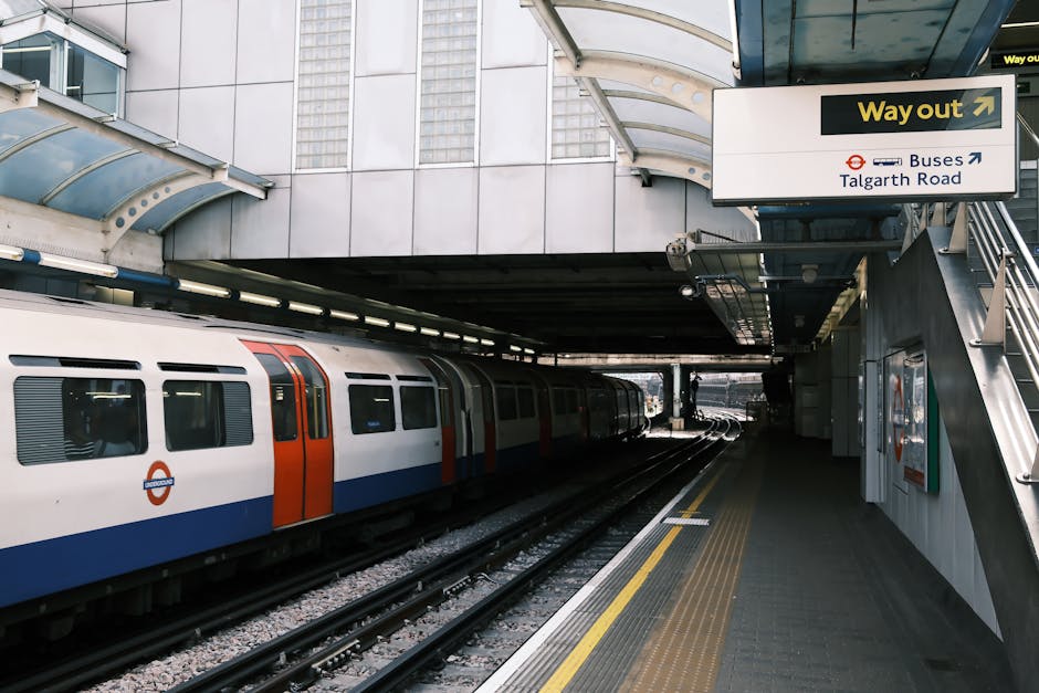 A modern London underground train with a white, blue, and orange exterior is stationary at an underground station platform, which is made of concrete with yellow tactile paving along the edge for safety. Overhead, a curved glass canopy provides partial shelter, and an overhead sign indicates directions to buses towards Talgate Road and an exit route. To the right, a staircase with metal railings leads up to an exit or connecting walkway. The station environment features metallic and glass elements, with lighting integrated into the ceiling. This setting represents a typical urban transit environment used by residents and workers in Hammersmith, where [COMPANY_NAME] provides house removals and furniture transport services, ensuring efficient planning for home relocation or moving logistics involving transport via public transit and connecting facilities.