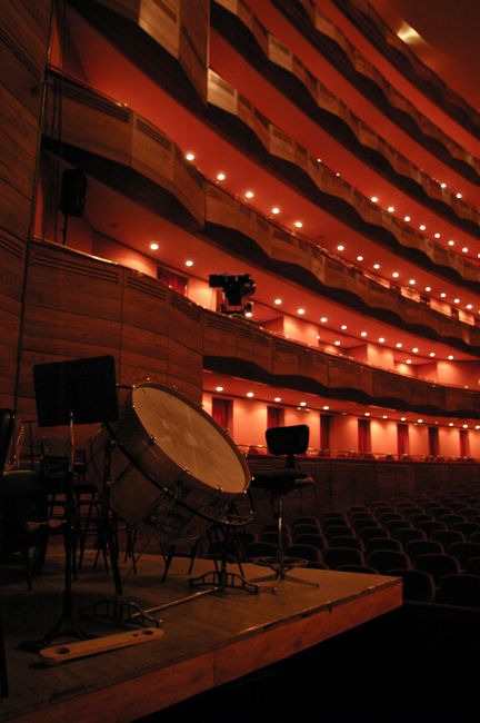 Inside the Lyric Hammersmith theatre, the image shows a stage area prepared for a music or theatrical production with several percussion instruments, including a large bass drum and a snare drum, placed on stands on a wooden platform. There are black chairs and stands nearby for musicians or performers. The auditorium features curved balconies with warm red lighting, creating a soft glow along the tiered seating levels. The theatre's interior walls are lined with wooden panels, enhancing the acoustics and aesthetics. The audience seating extends into the background with rows of upholstered chairs arranged in a semi-circular pattern facing the stage. The lighting suggests a quiet preparation phase before a stage performance, illustrating an environment suitable for stage staging moves or equipment setup that would be part of a house or venue relocation process, consistent with professional moving services like those offered by Hammersmith Movers.