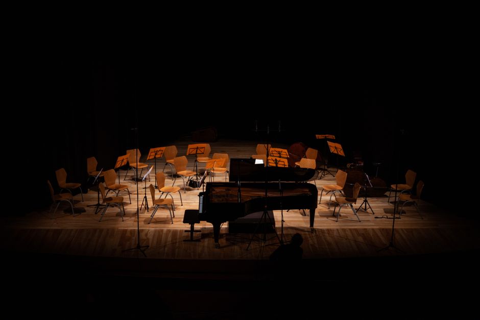 An empty stage inside a concert hall features rows of light brown chairs arranged in a semi-circle, each with metal legs and fabric upholstery, positioned behind music stands with sheet music. In front of the chairs, there is a grand piano with a closed lid, placed on a wooden floor, and several microphone stands are set up around the stage. The scene is dimly lit with focused lighting highlighting the chairs and the piano, while the background remains dark, indicating preparations for a musical performance or phase of staging moves. This setting relates to house removals and staging services offered by Hammersmith Movers, including furniture transport and careful packing procedures involved in home relocation or musical equipment staging moves as referenced in the Lyric Hammersmith instrument removals and staging moves page.