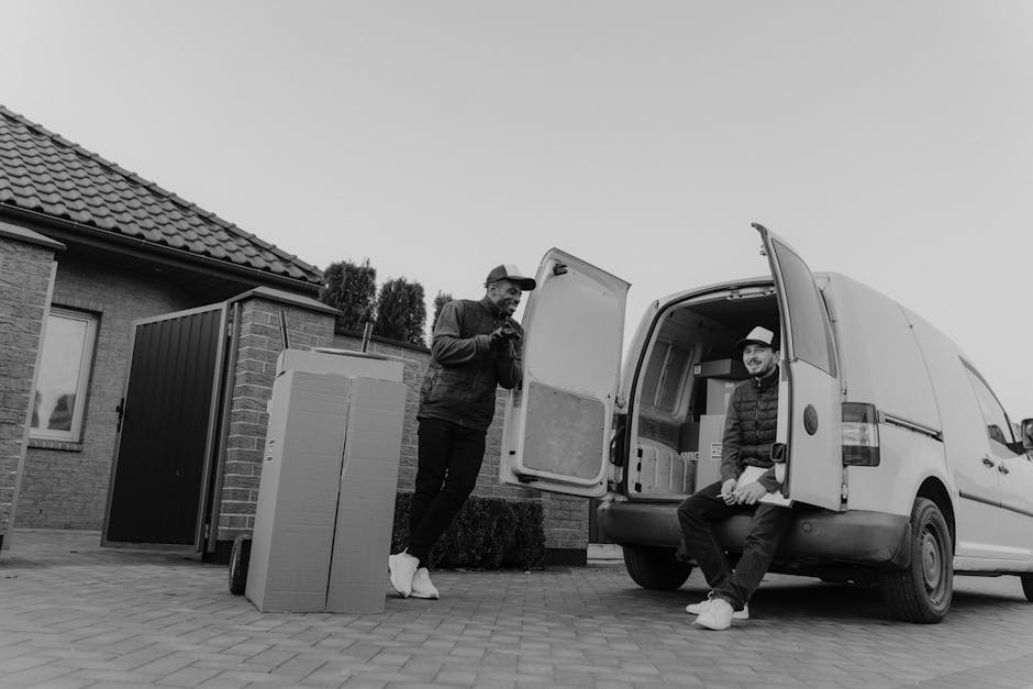 Two men involved in a home relocation process near a residential property with a tiled roof and brickwork. One man is standing outside on paved ground, leaning against a cardboard box, while the other is seated on the open rear door of a white moving van, wearing a hat and jacket, both appearing to be taking a break or coordinating the packing and loading process. The open van is partially filled with boxed items and packing materials, including plastic-wrapped furniture and other packing supplies. Nearby, a large black and white plastic crate with wheels is positioned on the pavement, indicating the use of moving equipment such as trolleys. The scene is set in daylight with clear visibility, and the surroundings include a small garden hedge, contributing to a typical house removal environment. Hammersmith Movers handle this task, illustrating their house removals and furniture transport services in W6, Hammersmith, as they assist with packing, loading, and logistics of the move.