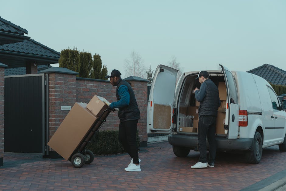 Two removal workers from Hammersmith Movers are engaged in the home relocation process outside a residential property. One worker, dressed in a blue and black jacket with a cap, is pushing a hand truck loaded with a large cardboard box towards the open rear doors of a white moving van parked on a paved driveway. The other worker, wearing a black jacket and cap, is inside the van, organizing and packing additional boxes and packing materials such as cardboard cartons and wrapped furniture. The van's interior is partly visible, showing more boxes and packing supplies, with the rear doors swung open. The scene is set outdoors in a suburban area with brick walls, a black gate, and neighboring houses with tiled roofs in the background, under overcast or early evening lighting. This image captures the logistics and loading process involved in a professional furniture transport and packing service for house removals in the Hammersmith W6 area.