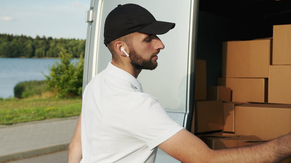 Two men involved in a home relocation process near a residential property with a tiled roof and brickwork. One man is standing outside on paved ground, leaning against a cardboard box, while the other is seated on the open rear door of a white moving van, wearing a hat and jacket, both appearing to be taking a break or coordinating the packing and loading process. The open van is partially filled with boxed items and packing materials, including plastic-wrapped furniture and other packing supplies. Nearby, a large black and white plastic crate with wheels is positioned on the pavement, indicating the use of moving equipment such as trolleys. The scene is set in daylight with clear visibility, and the surroundings include a small garden hedge, contributing to a typical house removal environment. Hammersmith Movers handle this task, illustrating their house removals and furniture transport services in W6, Hammersmith, as they assist with packing, loading, and logistics of the move.