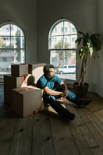 Inside a residential property, a man with curly dark hair and medium skin tone is sitting on the wooden floor next to a stack of cardboard boxes, some sealed with red packing tape. The boxes are of various sizes, arranged against a plain white wall, near two large arched windows that provide natural daylight. Outside the windows, a street with parked cars and buildings is visible. The man appears to be taking a break during a home relocation or packing process, with no other furniture or items present in the room. To his right, there is a tall potted plant with broad green leaves, adding a natural element to the space. The environment suggests a transit area for furniture transport and packing materials preparation, consistent with move or removals services, as provided by Hammersmith Movers.